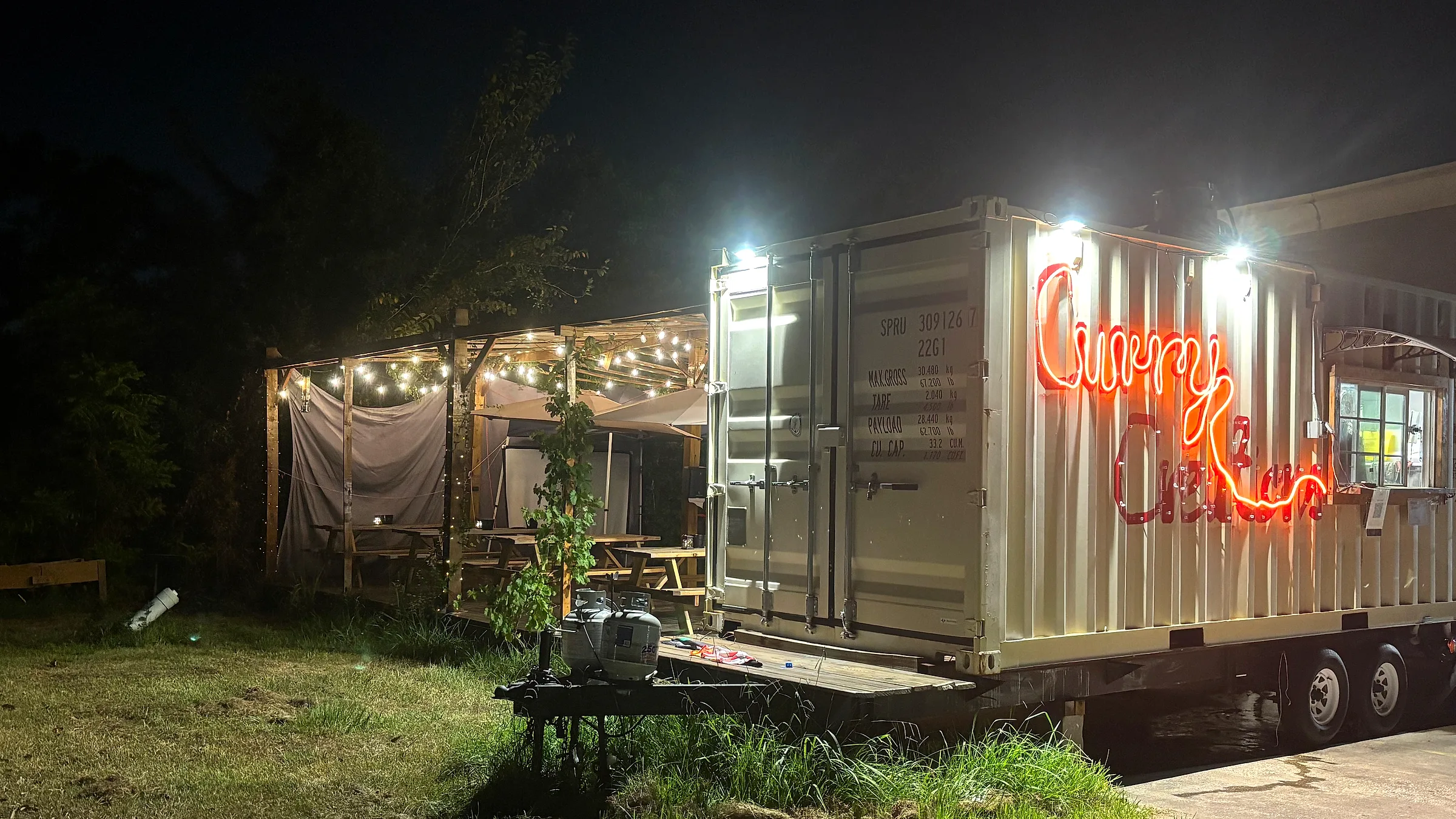 The Curry Creations trailer at night, its red neon sign lit against the container, with the string-lit patio alongside
